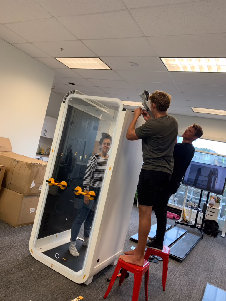 Two people assembling a single-person Bureau soundproof booth in an office, one standing on a red stool to fit the top frame while a third person stands inside the glass booth, with shipping boxes and a monitor showing the booth's 3D design nearby.