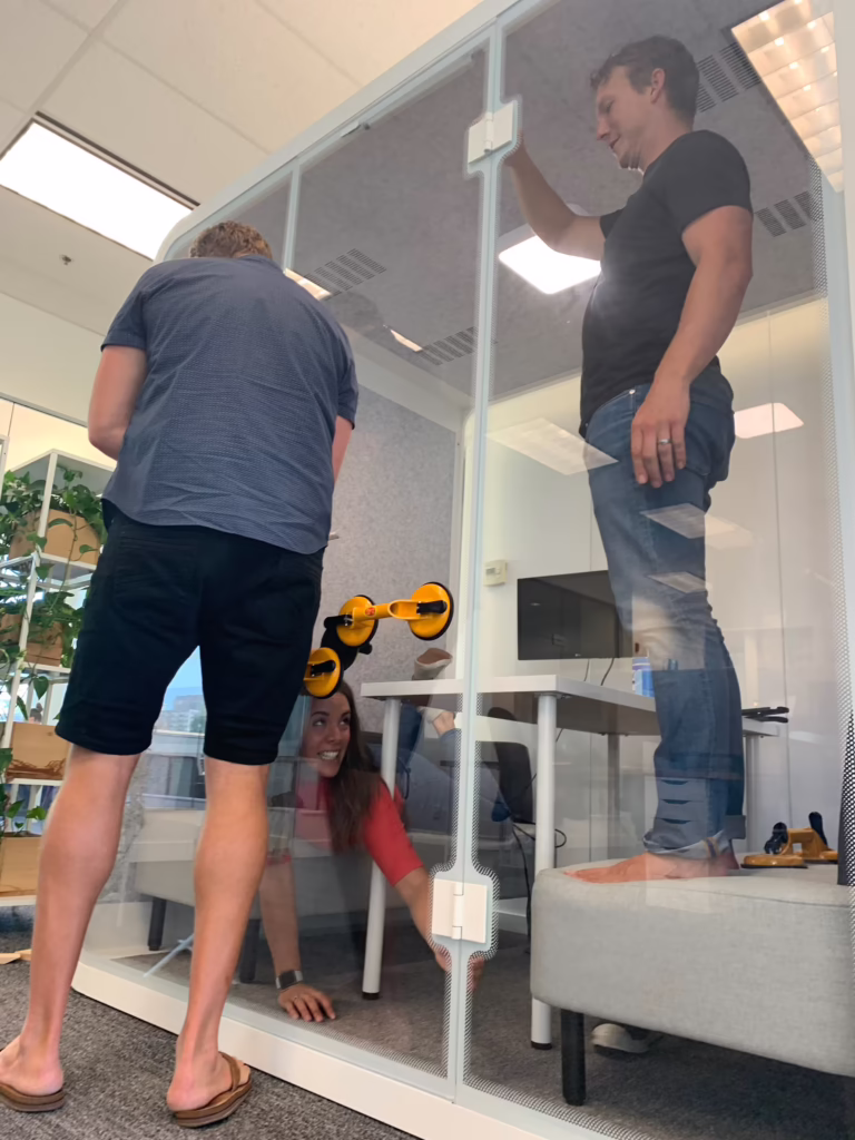 Three people installing a glass-walled Bureau office booth, one standing on the bench seat fitting the ceiling panel, another crouched at the base, and a third reaching up with suction-cup glass handles visible on the panel.