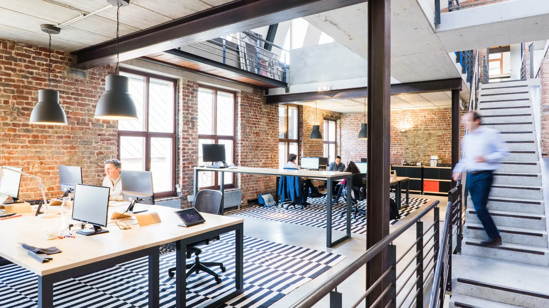 Modern open-plan office with exposed brick, open desks, and a staircase